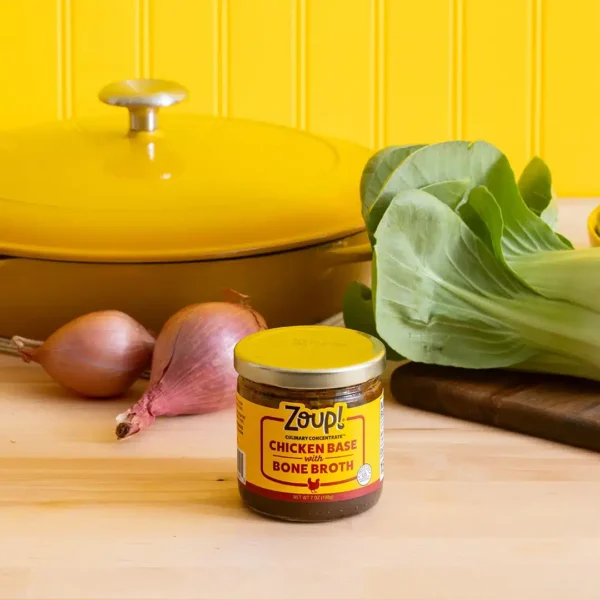 A jar of Zoup Chicken Base with Bone Broth on a light wood countertop with bok choy, shallots, and a yellow Dutch oven in the background.