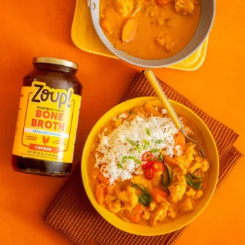 Overhead shot of a jar of Zoup Reduced Sodium Chicken Bone Broth laying flat next to a bowl of Thai coconut curry with rice, carrots, cauliflower, spinach, and red chili, with a pan of curry above.