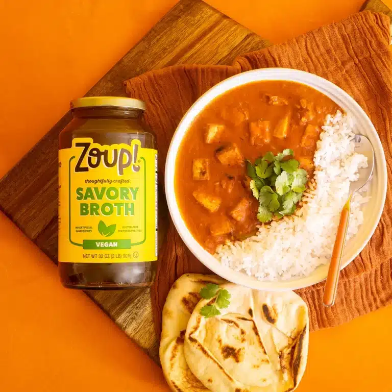 Overhead shot of a jar of Zoup Vegan Savory Broth next to a bowl of tofu Tikka Masala with white rice and cilantro, served with naan on a dark wooden board.