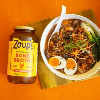 A jar of Zoup Spicy Chicken Bone Broth lays flat next to a bowl of ramen topped with soft-boiled eggs, mushrooms, bok choy, and shredded vegetables on an orange background.