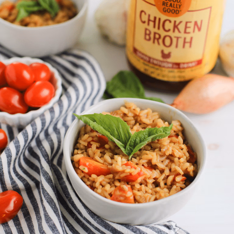 Creamy risotto rice in a white bowl with pieces of tomato, topped with basil leaves, placed in front of a jar of Zoup Chicken Broth.