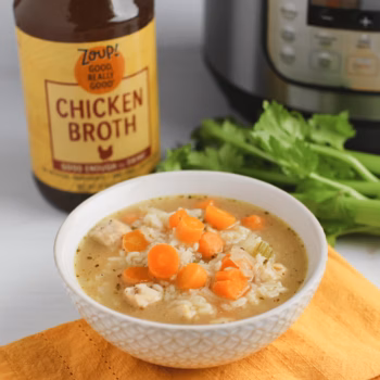 A white bowl of brothy chicken soup with rice, carrots celery and onion placed in front of an Instant Pot and jar of Zoup Chicken Broth.