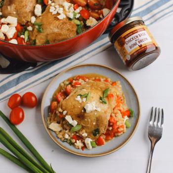 Cooked chicken thigh served over pearl couscous with diced tomatoes, green onions, and feta on a blue plate, with a red Dutch oven and Zoup Chicken Base Concentrate nearby.