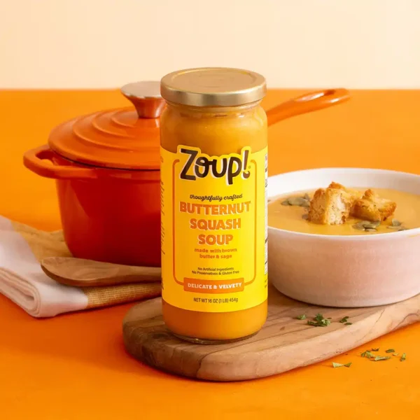 A jar of Zoup Butternut Squash Soup on a wooden cutting board beside a bowl of soup topped with croutons and sunflower seeds, with an orange Dutch oven in the background.