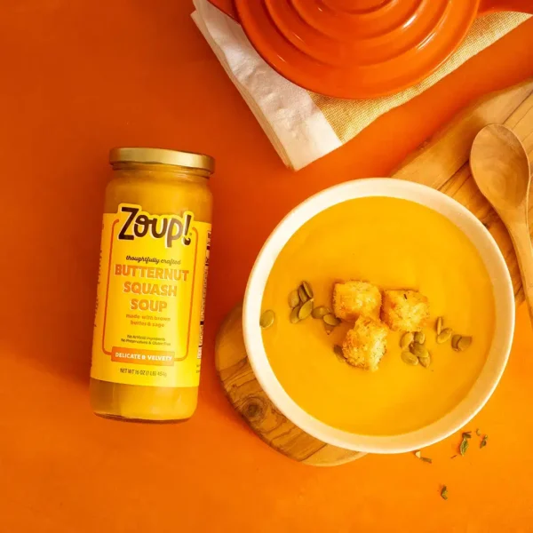 Overhead shot of a jar of Zoup Butternut Squash Soup beside a bowl of soup topped with croutons and pumpkin seeds, on an orange background with wooden utensils.