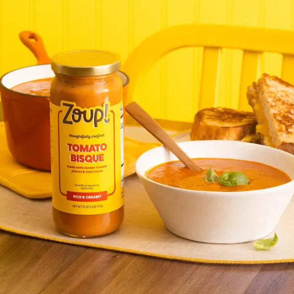 A jar of Tomato Bisque beside a bowl of soup garnished with basil, a wooden spoon, grilled cheese sandwiches, and a red pot of bisque in the background.