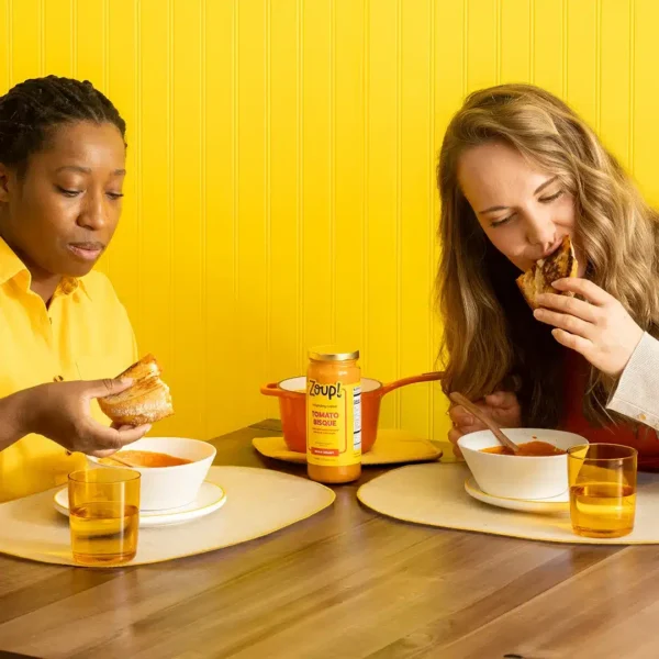 Two women enjoying grilled cheese and bowls of Zoup Tomato Bisque at a table with a jar of soup and an orange pot in the center.