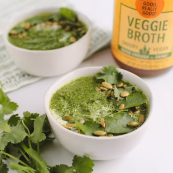Two white bowls of blended green soup topped with cilantro and pumpkin seeds, in front of a jar of Zoup Veggie Broth.