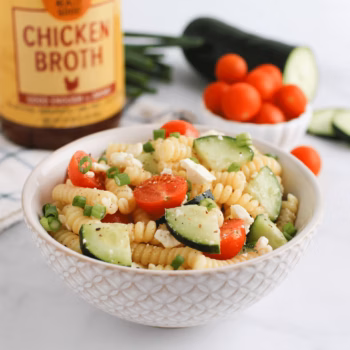 Pasta salad with cucumbers, cherry tomatoes, feta, and green onions in a white textured bowl, with Zoup Chicken Broth and fresh veggies in the background.