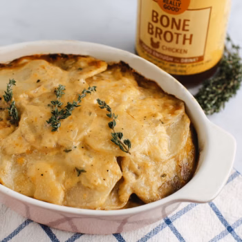 A white casserole dish filled with creamy sliced potatoes topped with melted cheese and topped with sprigs of thyme, in front of a jar of Zoup Chicken Bone Broth.