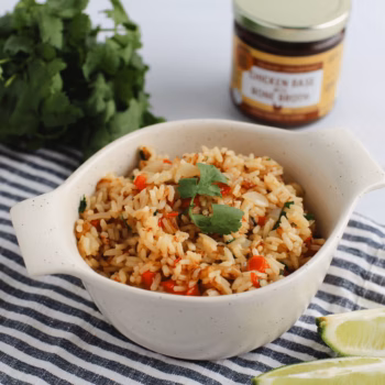 A bowl of seasoned rice with chopped red peppers and cilantro, set on a striped cloth with lime wedges and a jar of Zoup chicken base Concentrate in the background.