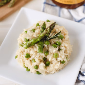 Creamy risotto with green peas, topped with grilled asparagus and a sprig of rosemary, served on a square white plate.