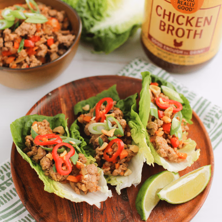Three lettuce wraps filled with seasoned ground turkey, sliced red peppers, green onions, and chopped peanuts, served with lime wedges and placed beside a jar of Zoup chicken broth.