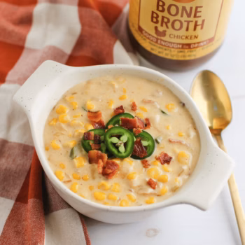 Creamy corn chowder in a white bowl topped with jalapeño slices and crispy bacon bits. A jar of Zoup Chicken Bone Broth sits in the background.