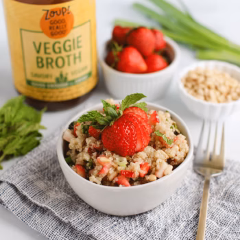 White bowl filled with quinoa salad topped with chopped strawberries, fresh mint, and pine nuts. A jar of Zoup Veggie Broth and bowl of strawberries are in the background.