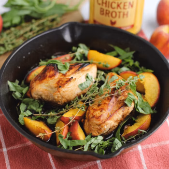 Cast iron skillet with seared chicken breasts, sliced peaches, and fresh herbs. A jar of Zoup Chicken Broth sits in the background on a red towel.