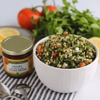 White bowl filled with fresh tabouli made of bulgur, parsley, tomato, and onion. Surrounded by lemons, parsley, and a jar of Zoup Vegan Broth Concentrate.
