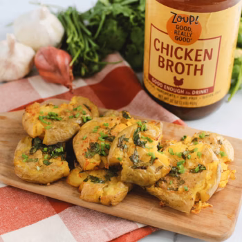 Baked smashed potatoes stacked on a wooden cutting board and sprinkled with chives, with a jar of Zoup Chicken broth in the background