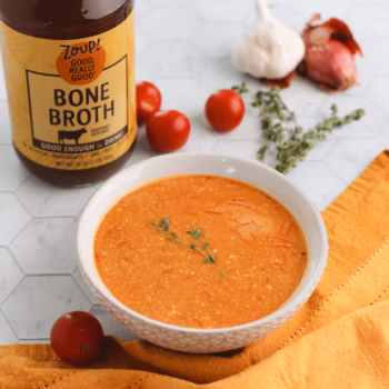 A bowl of baked tomato feta soup topped with fresh thyme, surrounded by garlic, shallots, cherry tomatoes, and a jar of Zoup Beef Bone Broth on a hex tile surface.