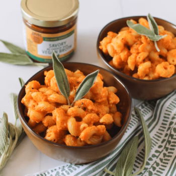 Bowls of creamy one-pot pumpkin pasta garnished with fresh sage leaves, surrounded by loose sage and a jar of Zoup! Savory Vegan Broth on a white surface.