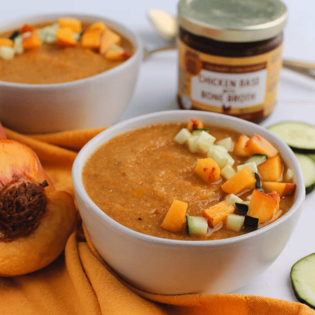 Bowl of peach gazpacho topped with diced peaches and cucumber, with fresh peaches, cucumber slices, and a jar of Zoup Chicken Bone Broth Base nearby.
