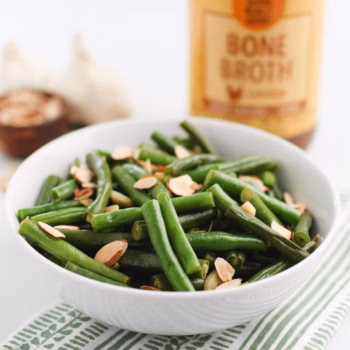 A bowl of green beans topped with toasted sliced almonds, with garlic cloves and a jar of Zoup Chicken Bone Broth blurred in the background.