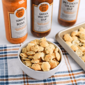Bowl of oyster crackers tossed with ranch seasoning, next to several jars of Zoup soup next to a baking tray.