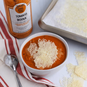 Tomato bisque served in a white bowl and topped with homemade parmesan crisps, with a bottle of Zoup Tomato Bisque and a baking tray of crisps in the background.