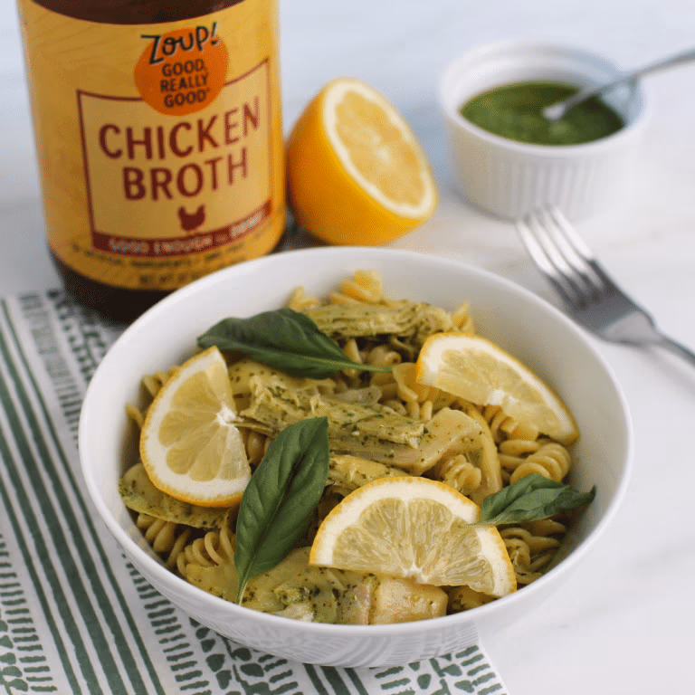 A bowl of pesto artichoke pasta topped with lemon slices and fresh basil, with Zoup Chicken Broth, pesto sauce, and a halved lemon in the background.