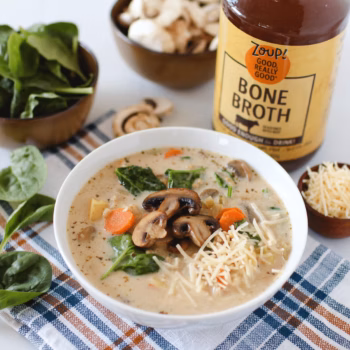 Bowl of creamy mushroom and rice soup with carrots, spinach, and shredded parmesan. A jar of Zoup Bone Broth and fresh veggies are placed around the bowl.