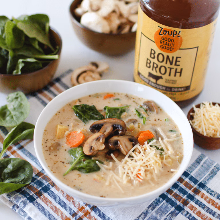 Bowl of creamy mushroom and rice soup with carrots, spinach, and shredded parmesan. A jar of Zoup Bone Broth and fresh veggies are placed around the bowl.