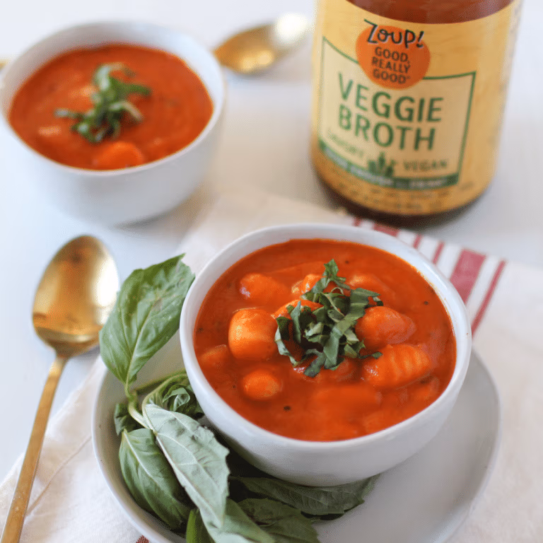 Bowls of vegan tomato soup with pillowy gnocchi, topped with fresh basil. A jar of Zoup! Veggie Broth and a bunch of basil rest beside the soup.