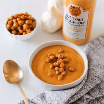 A bowl of butternut squash soup topped with crispy chickpeas, set next to a jar of Zoup Butternut Squash Soup, garlic bulbs, and a textured napkin.