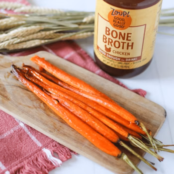 A stack of whole peeled, glazed roasted carrots arranged on a wooden board with a red cloth underneath. A jar of Zoup! Chicken Bone Broth and wheat stalks are in the background.