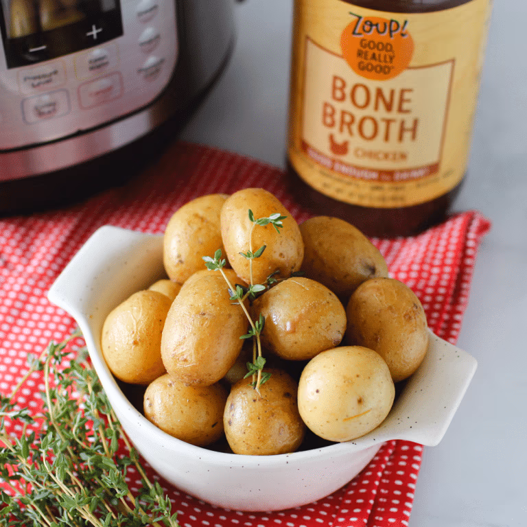 A bowl of baby potatoes garnished with thyme, set on a red towel beside an Instant Pot and a jar of Zoup! Chicken Bone Broth