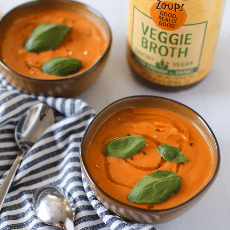 Bowl of creamy pumpkin sweet potato soup topped with fresh basil leaves. A jar of Zoup Veggie Broth and a striped towel with spoons sit beside the bowls.