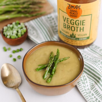 a bowl of creamy asparagus pea soup topped with whole peas and herbs. Fresh asparagus, lemon slices, and a jar of Zoup broth are placed in the background.