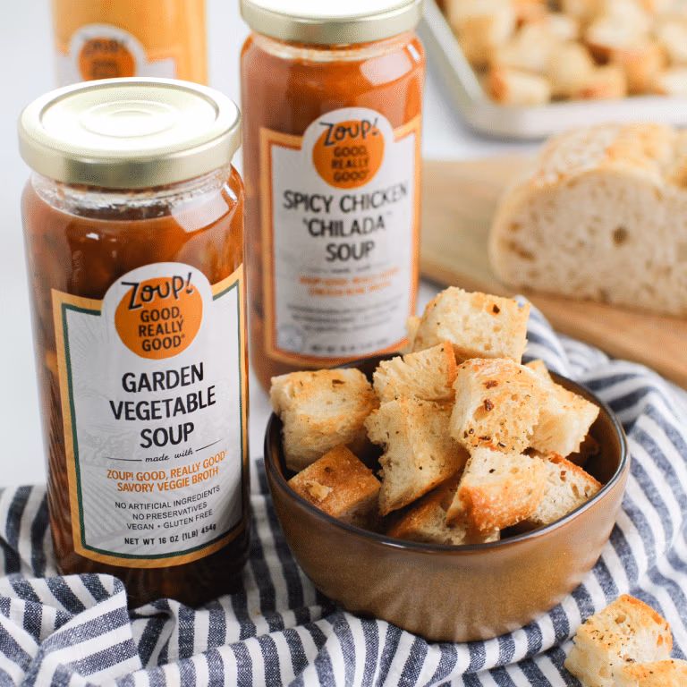 A small bowl of croutons sits beside several varieties of Zoup soup jars on top of a striped cloth with a loaf of bread in the background