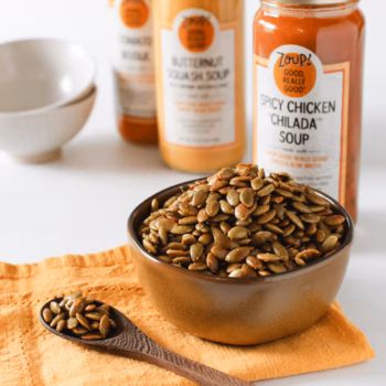 A wooden bowl filled with toasted sunflower seeds sits in front of several varieties of Zoup soup jars.