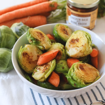 White bowl filled with roasted Brussels sprouts and carrots. Fresh carrots and a jar of Zoup Chicken Base Bone Broth Concentrate are in the background.