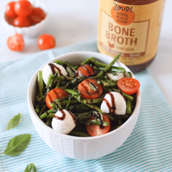 White bowl filled with asparagus caprese salad, featuring cherry tomatoes, mozzarella balls, and balsamic drizzle. Zoup Chicken Bone Broth in the background.
