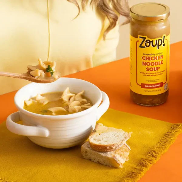 Close-up of a person holding a spoonful of Zoup Chicken Noodle Soup above a white bowl, with a jar of soup and bread on a yellow placemat.