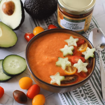 Bowl of creamy tomato gazpacho garnished with star-shaped cucumber slices and halved cherry tomatoes. Surrounded by fresh avocado, cucumber slices, colorful cherry tomatoes, and a jar of Zoup Savory Vegan culinary concentrate.