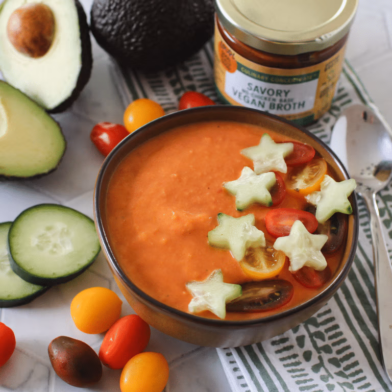 Bowl of creamy tomato gazpacho garnished with star-shaped cucumber slices and halved cherry tomatoes. Surrounded by fresh avocado, cucumber slices, colorful cherry tomatoes, and a jar of Zoup Savory Vegan culinary concentrate.