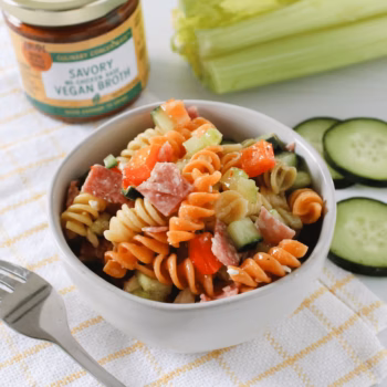 Tri-color rotini pasta salad with cucumber, bell pepper, and salami. Served in a white bowl with Zoup Vegan Broth concentrate nearby.