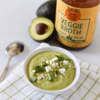 Bowl of creamy avocado gazpacho topped with crumbled cheese and fresh cilantro, placed beside a spoon, halved avocado, and a jar of Zoup Veggie Broth.