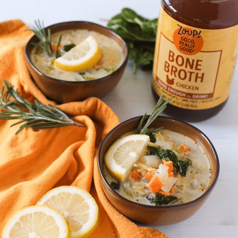 Bowls of creamy chicken orzo soup garnished with lemon slices and rosemary sprigs, placed alongside a jar of Zoup Chicken Bone Broth