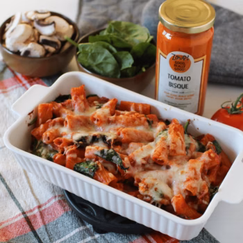 Baked rigatoni pasta with melted cheese, spinach, and mushrooms in a tomato bisque sauce, served in a white casserole dish next to a jar of Zoup Tomato Bisque, fresh spinach, and sliced mushrooms