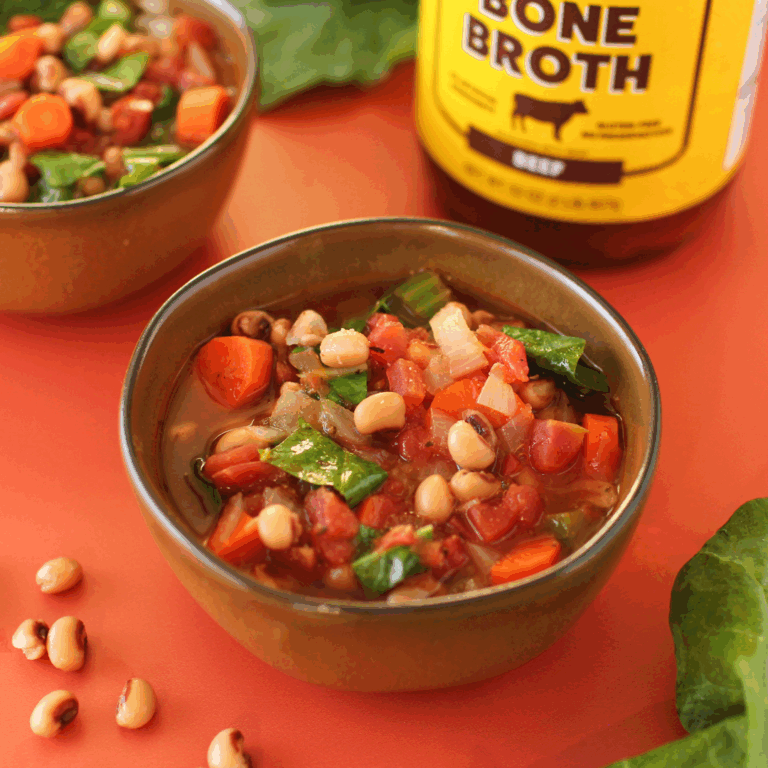 Bowls of black-eyed pea soup with spinach, carrots, and onions in a rich broth, served beside a jar of Zoup Beef Bone Broth and a gold spoon on a warm red table.