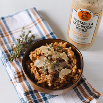 A bowl of creamy mushroom risotto topped with shaved parmesan and fresh thyme, set on a plaid kitchen towel next to a sprig of thyme and a jar of Zoup Portabella Mushroom Bisque
